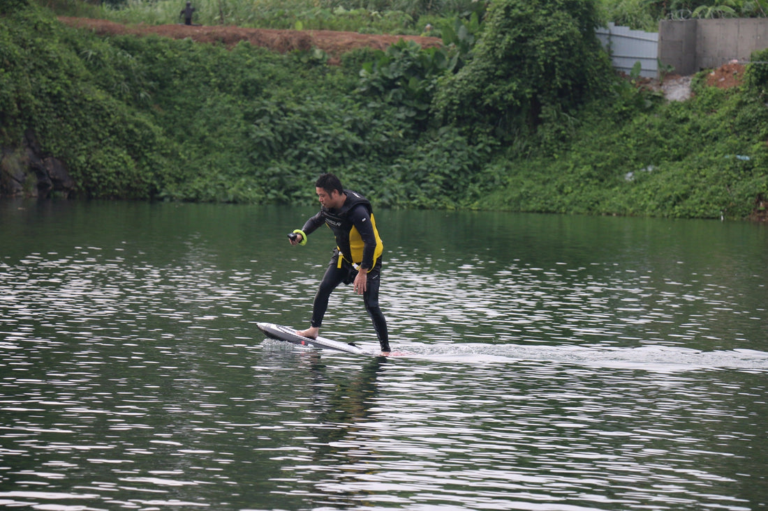 A man glides smoothly across calm waters on an electric hydrofoil surfboard, dressed in a black-and-yellow wetsuit.