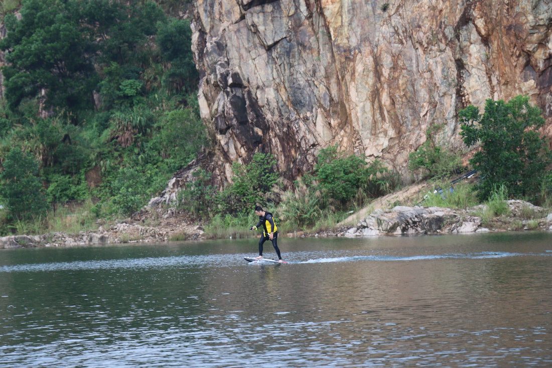 A person learning to ride an electric hydrofoil on a calm, mirror-like water surface, focusing on stability and control.